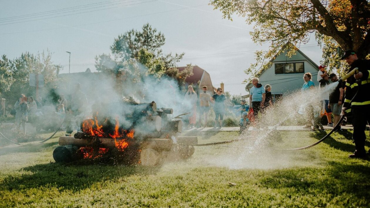 Firefighters spray water near a smoking outdoor fire as people watch